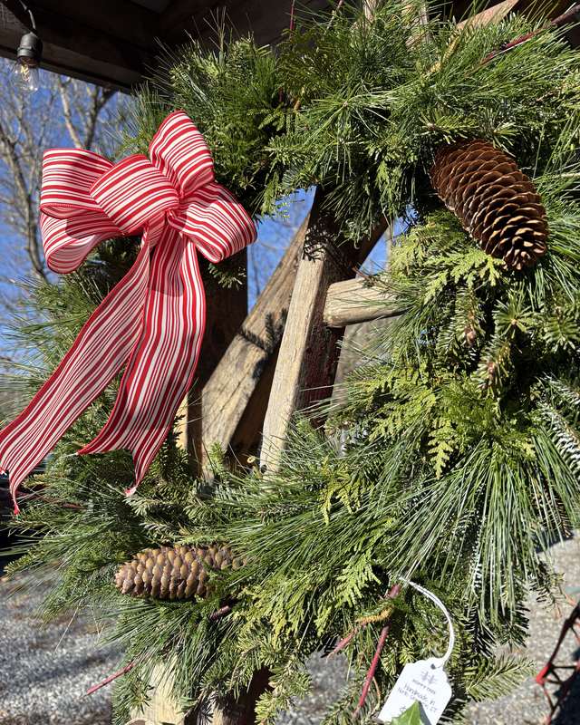 Fresh evergreen wreath decorated with a red-and-white striped bow and natural pinecones, hanging at the farm stand
